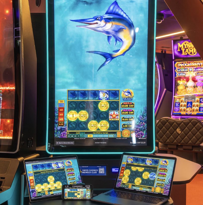 ReelLink on the Swiss Casinos floor: a slot cabinet running the Wazdan title Mighty Fish Blue Marlin, with the same game open on a tablet, a laptop and a phone in front of it